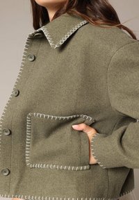 Veste en laine vert olive avec une coupe ample, coutures contrastantes blanches le long du col, des boutons et de la poche. Dotée d'une seule poche à rabat.