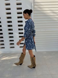Blå denimkjole med blomstermønster, innsvingt liv og splitt på siden, kombinert med beige semsket skinn cowboystøvler med frynser. Nøytral flislagt bakgrunn.