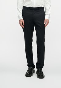 Melnas uzvalka bikses ar gludu audumu, taisnu piegriezumu, plakanu priekšpusi un jostu cilpiņām, savienotas ar melniem apaviem.