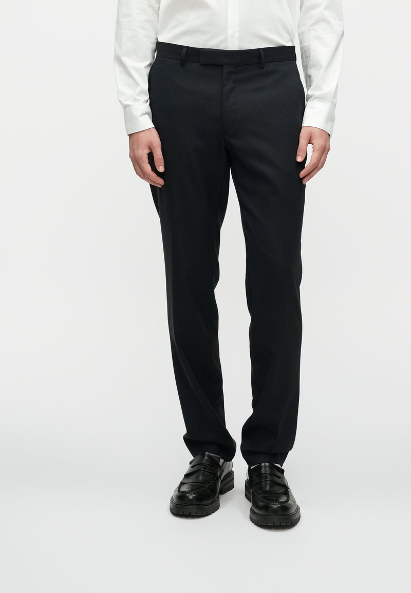 Melnas uzvalka bikses ar gludu audumu, taisnu piegriezumu, plakanu priekšpusi un jostu cilpiņām, savienotas ar melniem apaviem.