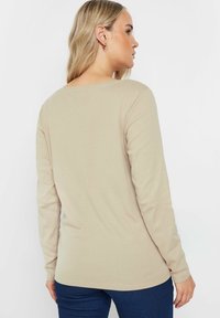 Camicia beige a maniche lunghe con scollatura rotonda, morbida al tatto e sagomata. Indossata con pantaloni in denim scuro, mostrando una schiena liscia.