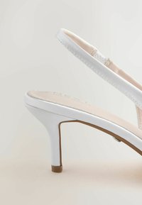Escarpin en cuir blanc à bride arrière avec une fine sangle et une semelle intérieure beige rembourrée sur un fond clair.