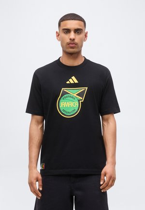 Νέος άνδρας φοράει μαύρο T-shirt με κίτρινο και πράσινο λογότυπο της Ποδοσφαιρικής Ομοσπονδίας της Τζαμάικας και σήμα Adidas μπροστά.