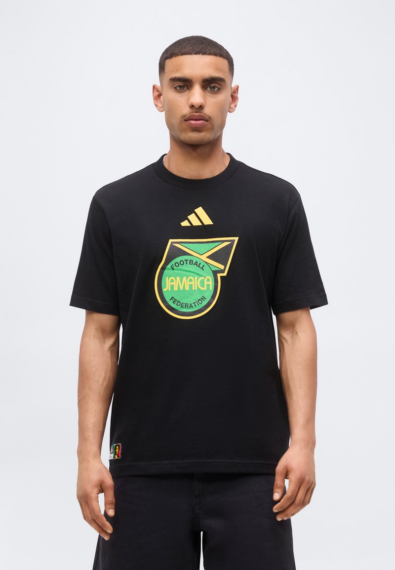 Νέος άνδρας φοράει μαύρο T-shirt με κίτρινο και πράσινο λογότυπο της Ποδοσφαιρικής Ομοσπονδίας της Τζαμάικας και σήμα Adidas μπροστά.