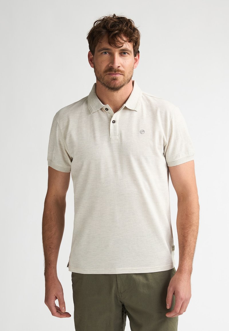 Homme aux cheveux bruns courts et à la barbe, portant un polo beige clair et un pantalon vert olive, debout devant un fond uni.