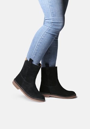 Person wearing light blue skinny jeans and black suede ankle boots standing on white background.