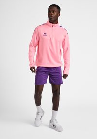 Maglietta sportiva rosa a maniche lunghe con colletto a mezza zip, con accenti viola sulle spalle, abbinata a pantaloni corti viola e scarpe da ginnastica bianche.