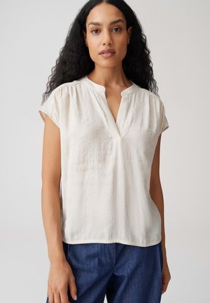 Femme aux longs cheveux bouclés portant un chemisier blanc à manches courtes et un pantalon bleu, debout devant un fond clair uni.