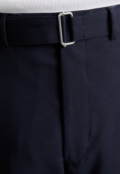 Pantalon bleu marine en tissu lisse, à devant plat, avec une boucle rectangulaire en métal argenté à la taille. Détails de couture soignés visibles.