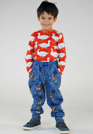 Glunderende jongen in een rood sweatshirt met witte eenden en een blauwe broek met rendieren die schaatsen, staand met zijn handen in zijn zakken tegen een effen achtergrond.