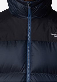 Doudoune bleu marine avec col montant, dotée d'une fermeture éclair intégrale et d'une texture en relief sur le tissu. Logo sur le haut à gauche.