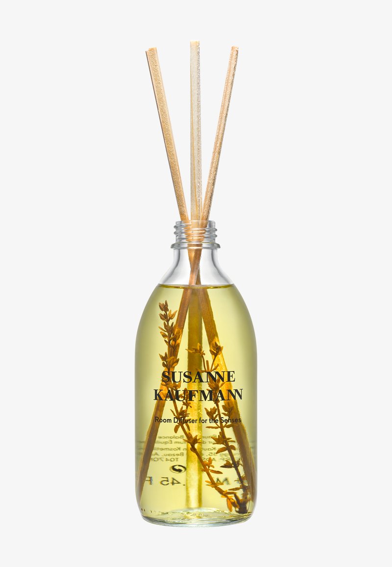 Flacon en verre transparent avec un liquide jaune, trois bâtonnets diffuseurs en rotin et des brins d’herbes séchées à l’intérieur, étiqueté Susanne Kaufmann diffuseur d’ambiance.