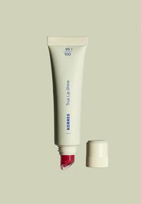 KORRES True Lip Shine dans un tube blanc, avec un texte bleu. Le bout du tube montre un produit rose brillant. Le capuchon est retiré, révélant une ouverture effilée.
