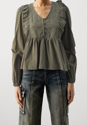 Mujer con blusa verde oliva con volantes de encaje y bordado floral, combinada con jeans cargo azul desvaído, posando sobre un fondo liso.