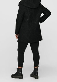 Abrigo negro con capucha, tejido texturizado y un corte ligeramente acampanado; usado sobre leggins negros y botas de tobillo con suela gruesa.