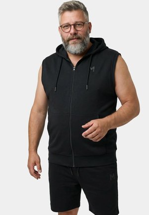 Mand med gråt skæg og briller iført sort ærmeløs hættetrøje og sorte shorts står foran en ensfarvet baggrund.