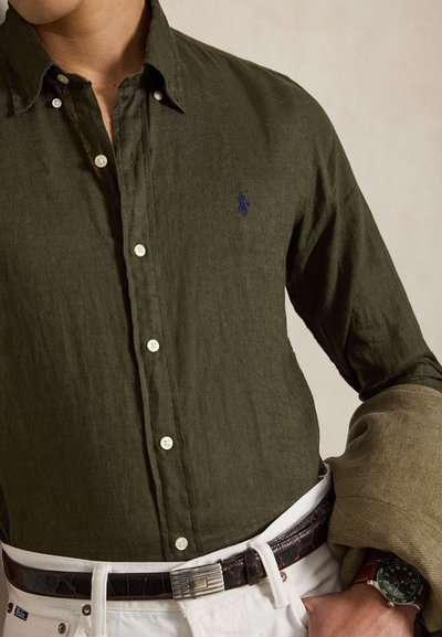 Férfi sötétzöld gombos ingben, kis hímzett logóval, fehér nadrág sötét övvel, kezében bézs dzsekit tart, karórát visel.