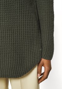 Texturovaný tmavě zelený pletený svetr s volným střihem a bocními rozparky. Má žebrované manžety a malou značkovou etiketu přišitou na lemu.
