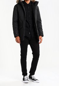 Parka nera con cappuccio foderato in pelliccia, dotata di due tasche frontali, abbinata a una camicia nera con bottoni, jeans neri e sneakers bianco e nere.