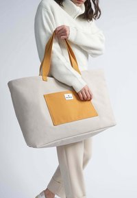 Große beigefarbene Tragetasche mit einer rechteckigen orangefarbenen Tasche. Verfügt über stabile gelbe Riemen und eine glatte Textur. Minimalistisches Design, praktisch für den täglichen Gebrauch.