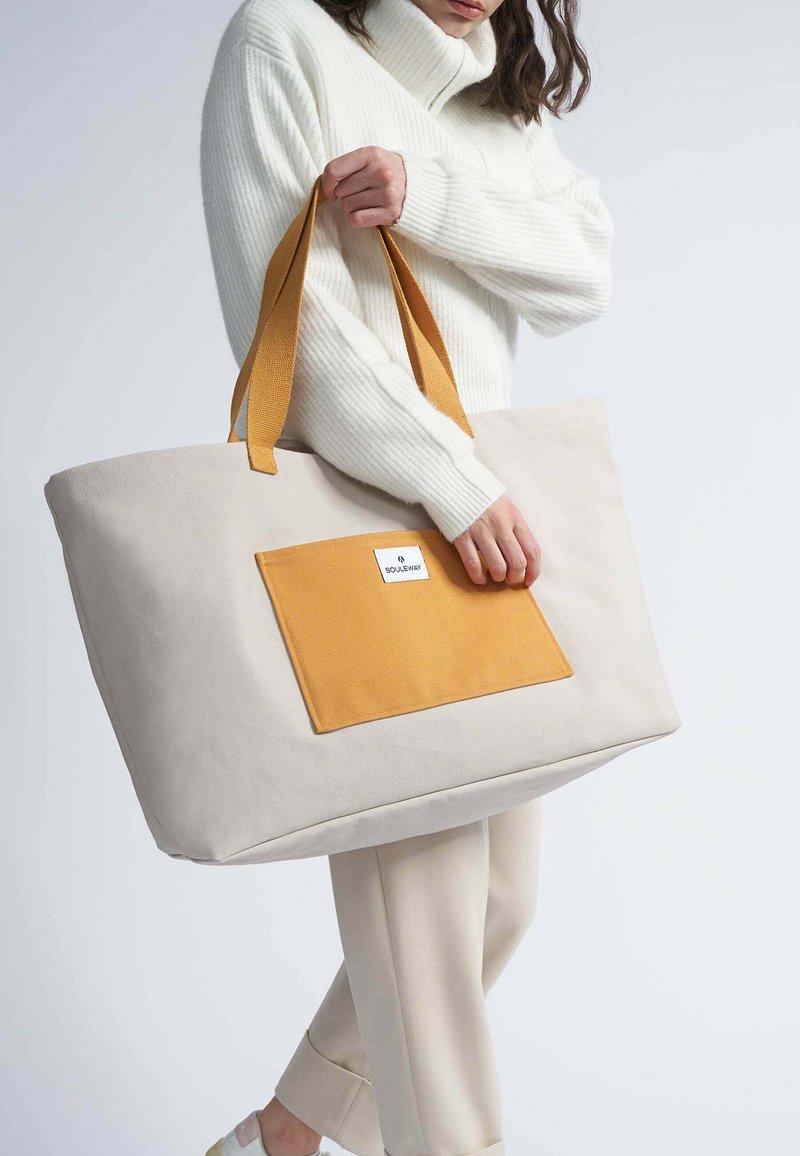 Große beigefarbene Tragetasche mit einer rechteckigen orangefarbenen Tasche. Verfügt über stabile gelbe Riemen und eine glatte Textur. Minimalistisches Design, praktisch für den täglichen Gebrauch.