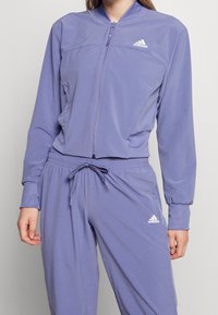 Kvinna iklädd en lila Adidas-träningsdräkt med en zip-up jacka och dragbandbyxor, båda med Adidas-logotypen på bröstet och benet.