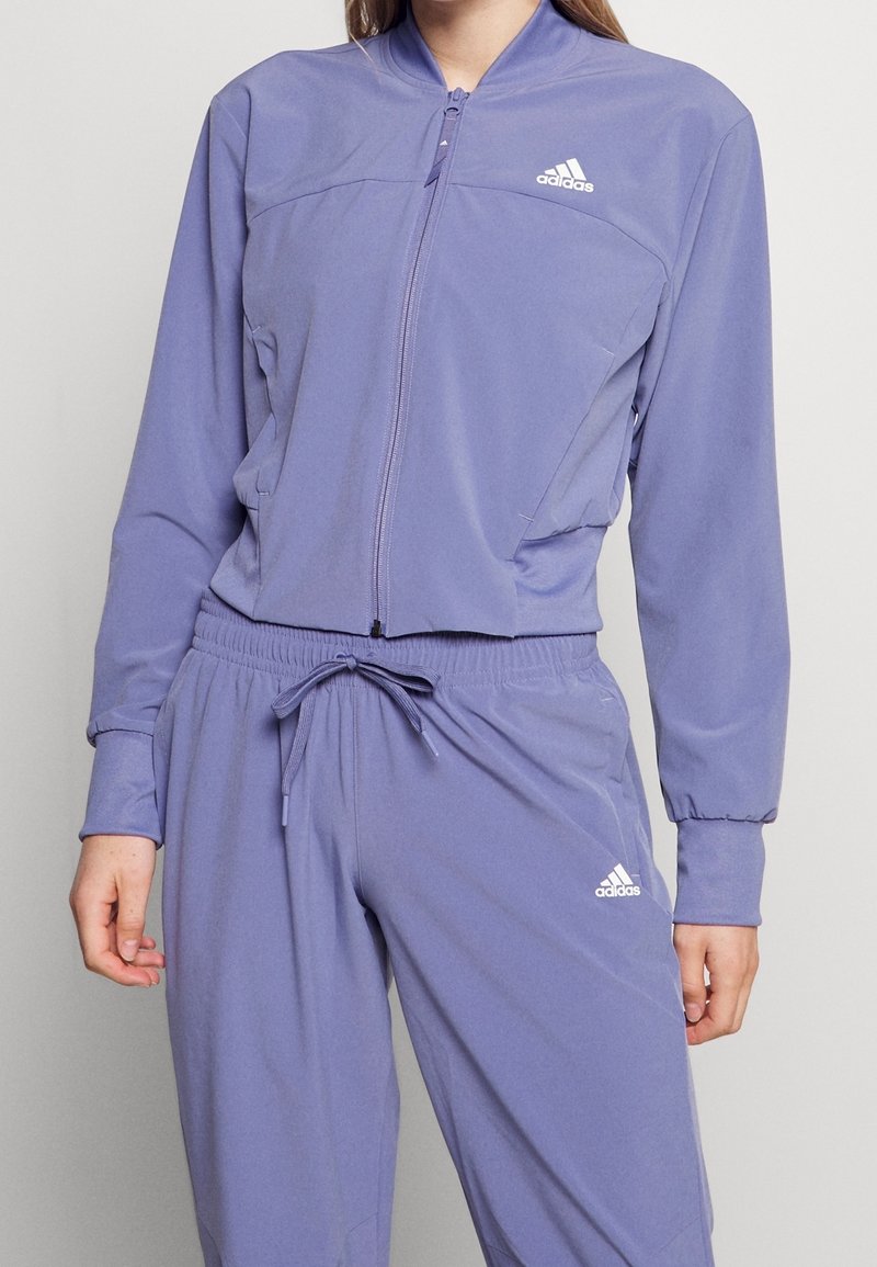 Kvinna iklädd en lila Adidas-träningsdräkt med en zip-up jacka och dragbandbyxor, båda med Adidas-logotypen på bröstet och benet.