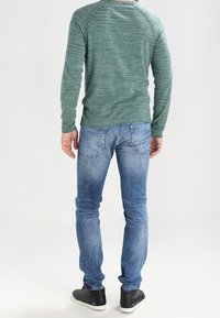 Grüner strukturierter Pullover mit Raglanärmeln, kombiniert mit ausgewaschenen blauen Jeans und schwarzen Sneakern. Das Bild zeigt eine Aufnahme von hinten.