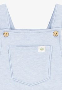 Pantalones de algodón azul con un bolsillo frontal cuadrado, dos botones de madera y una pequeña etiqueta cerca del bolsillo. Textura suave.