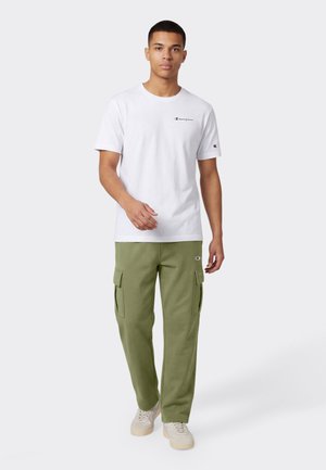 Młody mężczyzna idący, ubrany w białą koszulkę Champion, oliwkowo zielone spodnie cargo i białe tenisówki na jednolitym szarym tle.