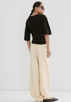 Vrouw met gevlochten haar en zonnebril, gekleed in een zwart topje, losse beige broek en zwarte puntige schoenen, staand tegen een effen witte achtergrond.