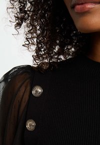 Eine Person mit lockigem Haar trägt ein schwarzes geripptes Oberteil mit transparenten Puffärmeln und zwei dekorativen goldenen Knöpfen an der Schulter.