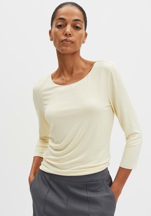 Helles gelbes, langärmeliges Oberteil mit rundem Halsausschnitt und weicher Textur, kombiniert mit dunkelgrauen Hosen. Zarte Drapierungsdetails an der Vorderseite.