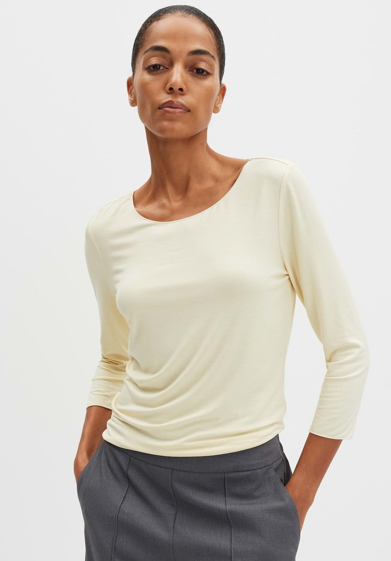 Lichtgele, langemouwen top met een ronde hals en een zachte textuur, gecombineerd met donkergrijze broeken. Subtiel drapeer-detail aan de voorkant.