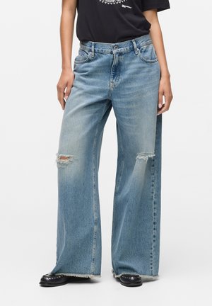 Lichtblauwe wijde spijkerbroek van denim met gescheurde knieën, rafelende zomen en een relaxte pasvorm, gedragen met een zwarte simpele graphic t-shirt.