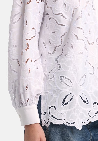 Weiße Bluse mit langen Ärmeln, verziert mit floraler Spitzenstickerei und mit wellenförmigem Saum, kombiniert mit blauen Denim-Jeans.