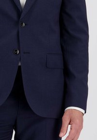 Man in een donkerblauwe blazer met twee knopen vastgemaakt, witte shirtmanchet zichtbaar, en bijpassende donkerblauwe broeken.