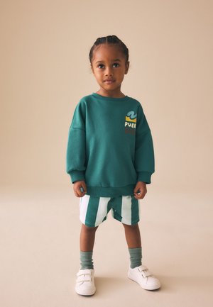 Fonott hajú gyerek zöld pulóvert, zöld-fehér csíkos rövidnadrágot, zöld zoknit és fehér tépőzáras tornacipőt visel, bézs háttér előtt áll.