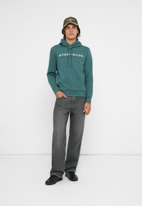 Felpa teal con logo frontale, cappuccio con cordino e polsini a costine. Abbinata a jeans grigi a vestibilità ampia e scarpe nere. Un cappello completa l'outfit.