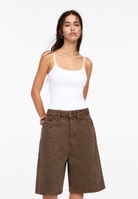 Bruine corduroy shorts met een hoge taille, voorzien van grote zijzakken, een knoopsluiting en een relaxte pasvorm. Gecombineerd met een witte tanktop.
