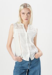 Weiße ärmellose Bluse mit strukturiertem Knötchenmuster, geknöpftem Vorderteil und Spitzenakzenten. Kombiniert mit hellblauen hochtaillierten Jeans.