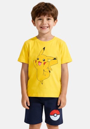 Garçon souriant portant un t-shirt jaune Pikachu et un short bleu marine avec un motif Poké Ball, debout sur un fond blanc.