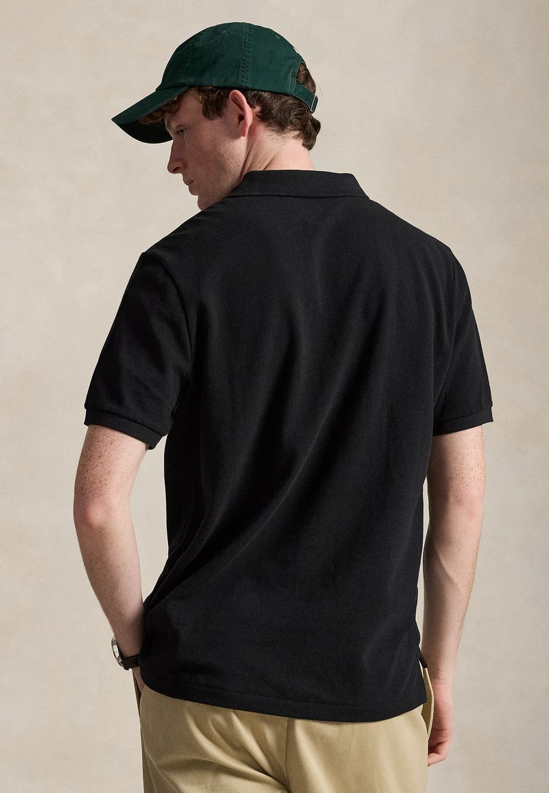 Mand iført sort poloshirt, beige bukser, grøn baseballkasket og ur, vendt let væk fra kameraet mod en ensfarvet baggrund.