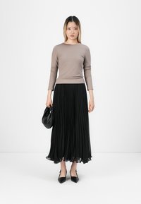 Egy nő áll, taupe színű, hosszú ujjú felsőt, fekete plisszírozott midi szoknyát és fekete hegyes orrú cipőt visel, fekete táska van a kezében, fehér háttér előtt.