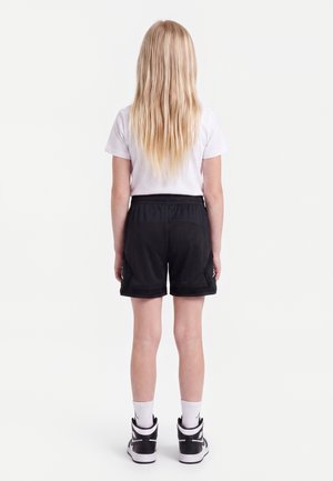 Enfant aux longs cheveux blonds, debout de dos, portant un t-shirt blanc, un short noir, des chaussettes blanches et des baskets noires et blanches sur un fond uni.