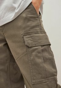 Pantaloni cargo in tessuto verde oliva con una texture liscia, dotati di molteplici tasche laterali e cuciture doppie per una maggiore resistenza.