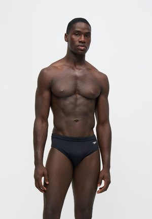 Muskuløs mand stående mod ensfarvet baggrund iført sort Speedo badebukser, vendt fremad med et neutralt udtryk.
