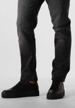 Zwarte sneakers met gestructureerde accenten en een gladde afwerking, gecombineerd met donkergrijze jeans. De schoenen hebben een dikke zool en een vetersluiting.