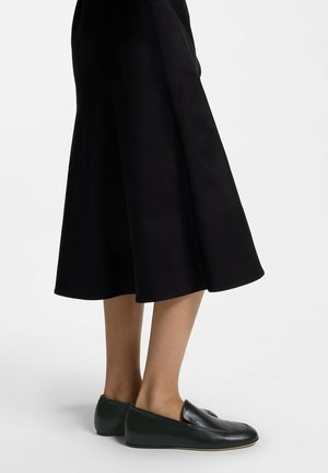 Beine, die schwarze Lederloafers und einen schwarzen Midirock tragen, vor weißem Hintergrund.