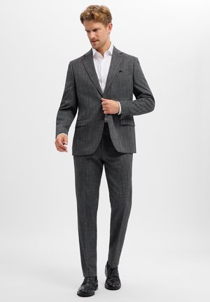 Man fastening button on dark gray pinstripe suit jacket worn with white shirt and black dress shoes on plain background.